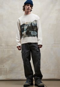 Krémová mikina s dlouhým rukávem s potiskem přírody, doplněná tmavými kalhotami typu cargo. Texturovaná, neformální obuv s modrými a šedými akcenty.