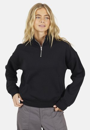 Kobieta o blond włosach, ubrana w czarny półgolf zapinany na zamek i szare spodnie, z rękami opartymi na biodrach, patrząca nieco na prawo od kamery.