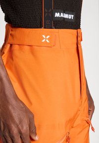 Κοντινό πλάνο ενός ατόμου που φορά φωτεινές πορτοκαλί παντελόνες με ρυθμιζόμενη μέση, επώνυμες τιράντες και μαύρο υφασμάτινο μπλουζάκι με το λάμδα "MAIMUT".