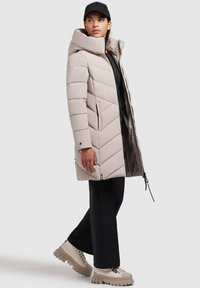 Polstret beige frakke med stor krave, lynlås og chevronmønster; sat sammen med sorte bukser og kraftige æggebene støvler.