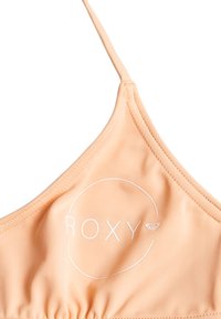 Persikofärgad bikiniöverdel med tunna band, mjuk textur och Roxy-logotyp i vitt inom en cirkulär design på framsidan.