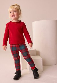 Top rojo de manga larga con detalle de volantes y acento atado, combinado con leggings a cuadros rojos y verdes; botas negras hasta el tobillo con detalles de borlas.