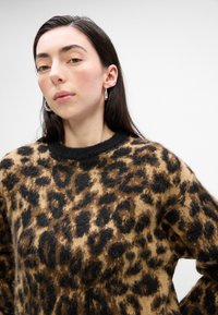 Svetr s leopardím vzorem v tanové a černé barvě, vyrobený z materiálu připomínajícího kožešinu, s černým žebrovaným límcem a příjemnou texturou. Jednoduchý design s volným střihem.