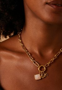 Gouden ketting met een ronde sluiting, een vierkante hanger en twee bedels: een schelp en een vogel. Glad metalen oppervlak.