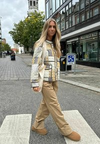 Jachetă matlasată cu modele geometrice în bej, gri și crem, asortată cu pantaloni khaki și papuci maronii, purtată pe o stradă din oraș.