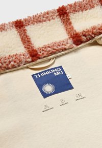 Cream-colored fabric with a fluffy, textured, reddish-brown collar. Features a blue label with logo and care icons for windbreaker, repellent, and breathability.