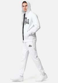 White zip-up hoodie with a black logo, paired with matching white joggers. Smooth fabric texture, relaxed fit, and front pockets.