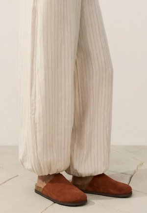 Beżowe luźne spodnie w cienkie prążki ze ściągaczami, noszone z brązowymi zamszowymi kapciami wsuwanymi na jasnej kafelkowej podłodze.