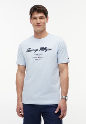 Uomo che indossa una t-shirt azzurro chiaro Tommy Hilfiger con logo e pantaloncini blu navy, in piedi su sfondo bianco, sorridente.