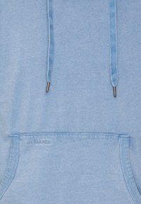 Světle modrá mikina s klokaní kapsou, stahovacími šňůrkami, kovovými konci a diskrétními logy na spodním okraji. Měkká textura a ležérní design.