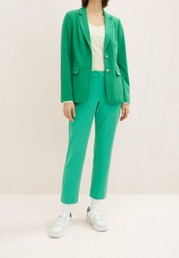 Femme portant un blazer vert vif et un pantalon assorti, avec un haut henley côtelé blanc et des baskets blanches, debout devant un fond uni.