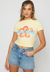 Camiseta amarilla corta con un gráfico de flor roja y naranja y "Surf Club" en texto azul, combinada con pantalones cortos de denim azul desgastados.