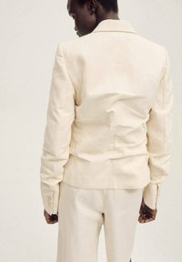 Beige maßgeschneiderte Blazer mit strukturiertem Design, ausgestattet mit einem Reverskragen und langen Ärmeln. Glattes, leichtes Material mit sichtbaren Nähten.