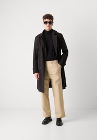 Manteau noir, col roulé noir, pantalon à taille élastique beige, chaussures noires et lunettes de soleil. Tissu lisse et coupe décontractée.