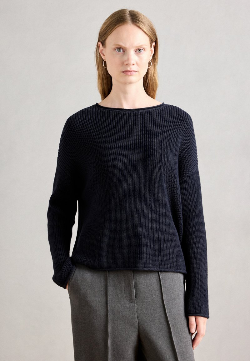 Maglione a coste blu navy con scollatura ampia e maniche lunghe, abbinato a pantaloni grigi sartoriali. Texture liscia, vestibilità casual.