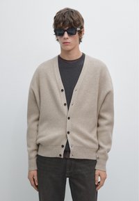 Beige geribbelde cardigan met knoopsluitingen, gedragen over een donkergrijs shirt, gecombineerd met zwarte rechthoekige zonnebril en donkere jeans.