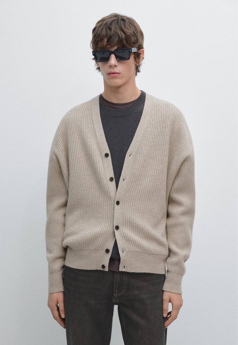 Beige geribbelde cardigan met knoopsluitingen, gedragen over een donkergrijs shirt, gecombineerd met zwarte rechthoekige zonnebril en donkere jeans.