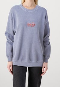Ljusblå sweatshirt gjord av mjukt material, med rund hals, långa ärmar och röd broderad text, samt ribbade muddar och nederdel.