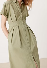 Olivově zelené šaty s knoflíky, límečkem, krátkými rukávy a plisovaným designem. Obsahují boční kapsy a přední rozparek. Hladká textilie.