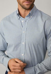 Camicia button-down a scacchi blu chiaro e bianca, con maniche lunghe, colletto e bottoni bianchi, che mette in mostra un tessuto di cotone testurizzato.