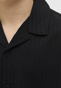 Camisa negra de textura con un patrón de rayas verticales, que presenta un cuello y cierre de botones. La tela parece suave y ligeramente arrugada.