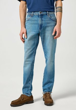 Man wearing light blue straight-leg jeans, brown lace-up shoes, and a navy blue shirt, standing against a plain background.