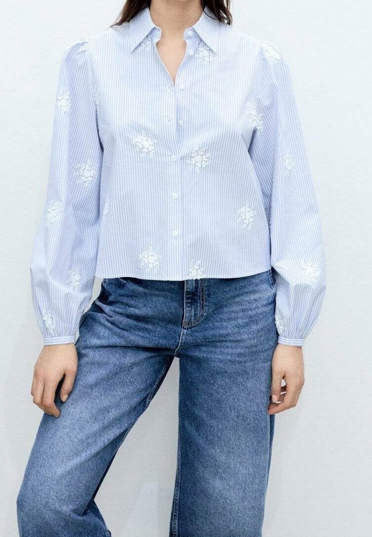 Femme portant un chemisier boutonné à rayures bleu clair avec une broderie florale blanche et un jean bleu, debout devant un fond clair.