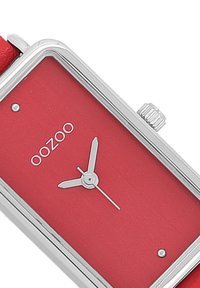 Montre rouge rectangulaire avec matériel en argent. Présente un cadran minimaliste avec trois aiguilles et le nom de la marque "oozoo" affiché de manière proéminente.
