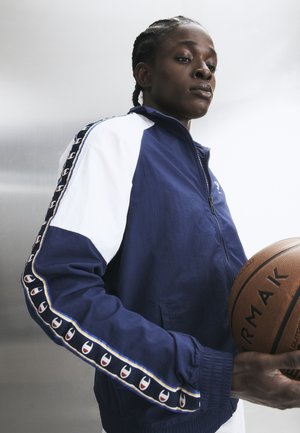 Idrettsutøver i blå og hvit jakke holder en basketball, ser fokusert og bestemt ut mot en nøytral bakgrunn.