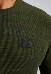 Grüner strukturierter Strickpullover mit einem schwarzen Etikett auf der Brust. Der Stoff hat ein geripptes Muster mit subtilen hellgrünen Sprenkeln.