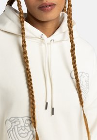 Cream hoodie featuring an embossed lion design, drawstrings with metal tips, and a relaxed fit. Smooth texture and minimalistic design.