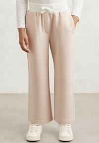 Šviesiai rožiniai treniruokliai su švelniu tekstūru, plačiomis kojomis ir balta elastinga juosta su mažyte mase. Kišenės ant priekinių šonų.