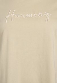 Beige bavlněné tričko s nápisem "Harmony" zdobeným imitací perel. Hladká struktura, krátké rukávy a volný střih.