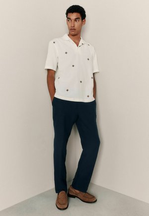 Weißes Kurzarmhemd mit kleinen braunen Rautenmuster, kombiniert mit marineblauen Hosen und braunen Wildleder-Loafers, vor einem neutralen Hintergrund stehend.