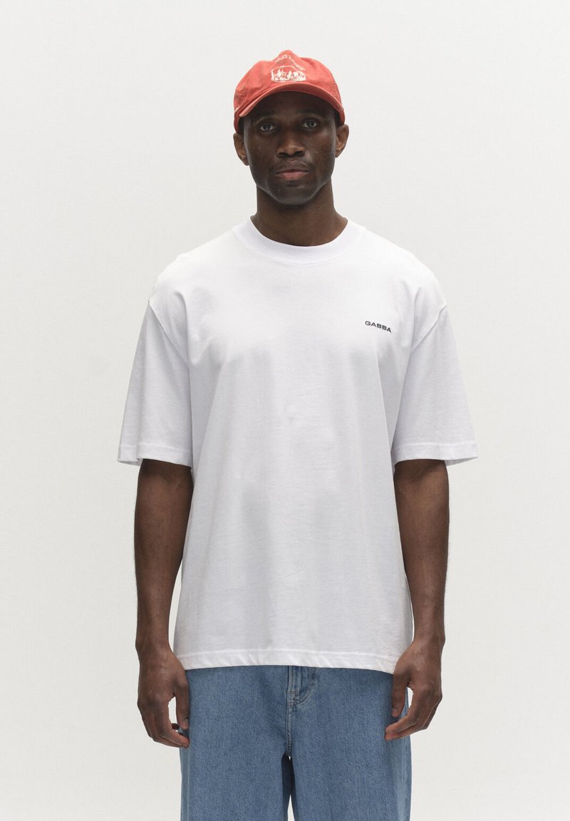Homme portant un T-shirt blanc uni avec le texte "GABBA", un jean bleu et une casquette rouge, debout devant un fond clair uni.