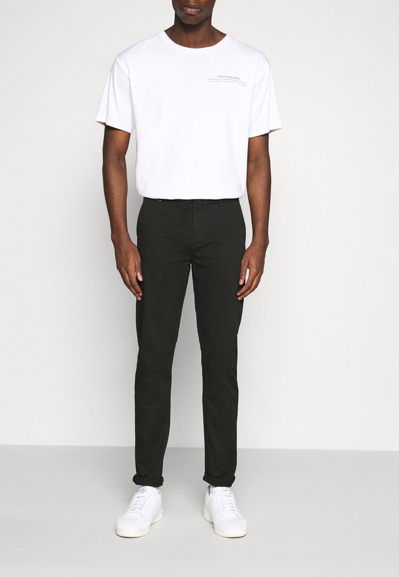 T-shirt blanche à manches courtes avec logo imprimé, associée à un pantalon slim green foncé et des baskets blanches, se tenant devant un arrière-plan neutre.