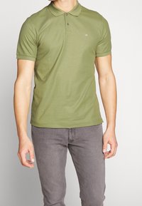 Polo vert olive avec une patte de boutonnage, manches courtes et petit logo sur la poitrine ; associé à un jean gris.