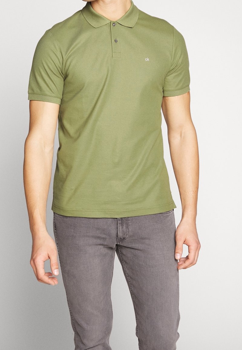 Polo vert olive avec une patte de boutonnage, manches courtes et petit logo sur la poitrine ; associé à un jean gris.