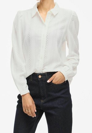 Mujer que lleva una blusa blanca de manga larga con ojales, metida dentro de unos vaqueros azul oscuro de cintura alta, con una mano en el bolsillo.