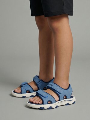 Kind trägt blaue sportliche Sandalen mit verstellbaren Riemen und weißen Sohlen, steht auf einer grauen Oberfläche.