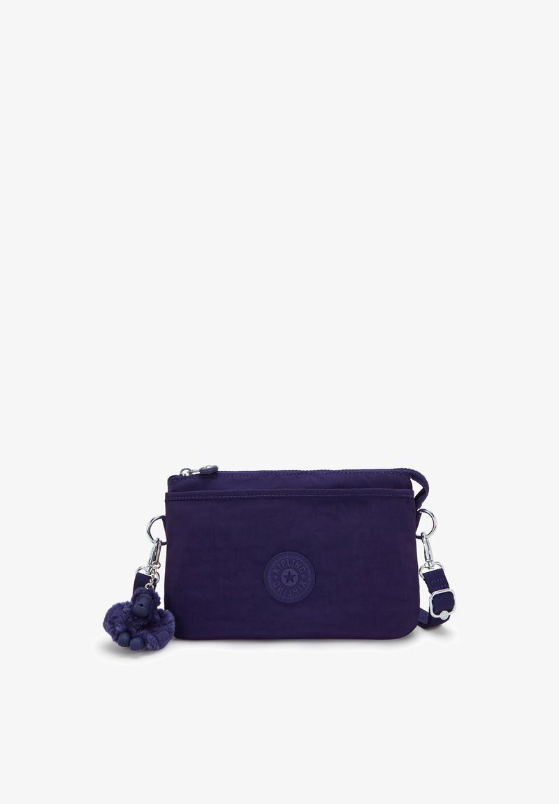 Piccola borsa a tracolla di colore viola scuro con tracolla regolabile, tasca frontale con zip, patch rotondo con logo e un portachiavi a forma di scimmia di peluche attaccato.