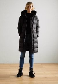 Cappotto piumino nero con cappuccio, caratterizzato da sezioni trapuntate, indossato su una maglietta nera e jeans blu. Il modello indossa sneakers alte nere.