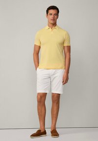 Keltainen poolopaita, jossa on kaulus, yhdistettynä valkoisiin shortseihin. Mallilla on ruskeat liukusandaalit. Minimalistinen muotoilu, ei kuoseja tai grafiikkaa.