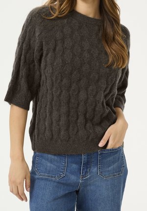 Femme portant un pull court à manches courtes en maille torsadée gris foncé, associé à un jean en denim bleu, la main posée dans la poche.