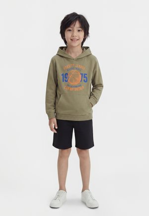 Garçon souriant portant un sweat à capuche de basketball vert olive, un short noir et des baskets blanches, debout avec une main dans la poche.
