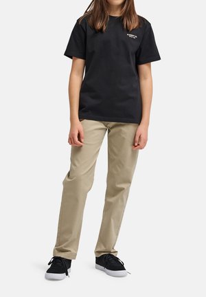 Jonge persoon die een zwart T-shirt met korte mouwen en beige broek draagt, staand met de handen ontspannen langs het lichaam, op een witte achtergrond.