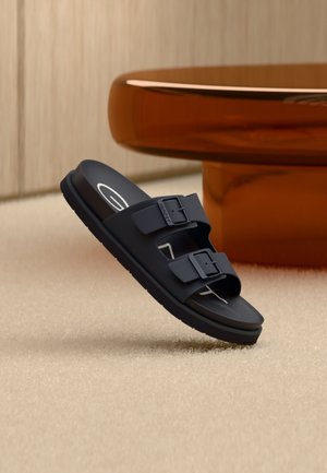 Μαύρο σανδάλι τύπου slip-on με δύο ρυθμιζόμενα λουριά με αγκράφες, που αιωρείται πάνω από μπεζ χαλί κοντά σε ένα καφέ στρογγυλό τραπέζι.