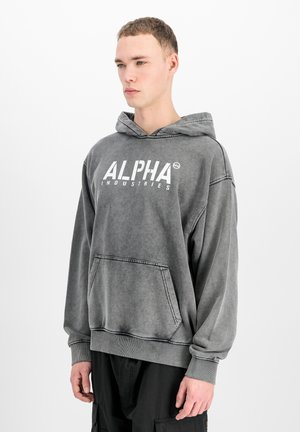 Mladi muškarac u izblijedjelom sivom Alpha Industries hoodicu s prednjim džepom stoji na jednobijelom zidu.