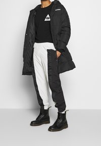 Svart puffjacka med huva, svart sweatshirt med en logotyp, svart och vita avsmalnande byxor med sidostråk, och svarta snörade kängor.