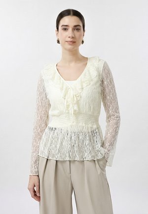 Femme aux cheveux foncés portant un chemisier en dentelle crème avec des volants et un pantalon plissé beige, debout sur fond blanc.